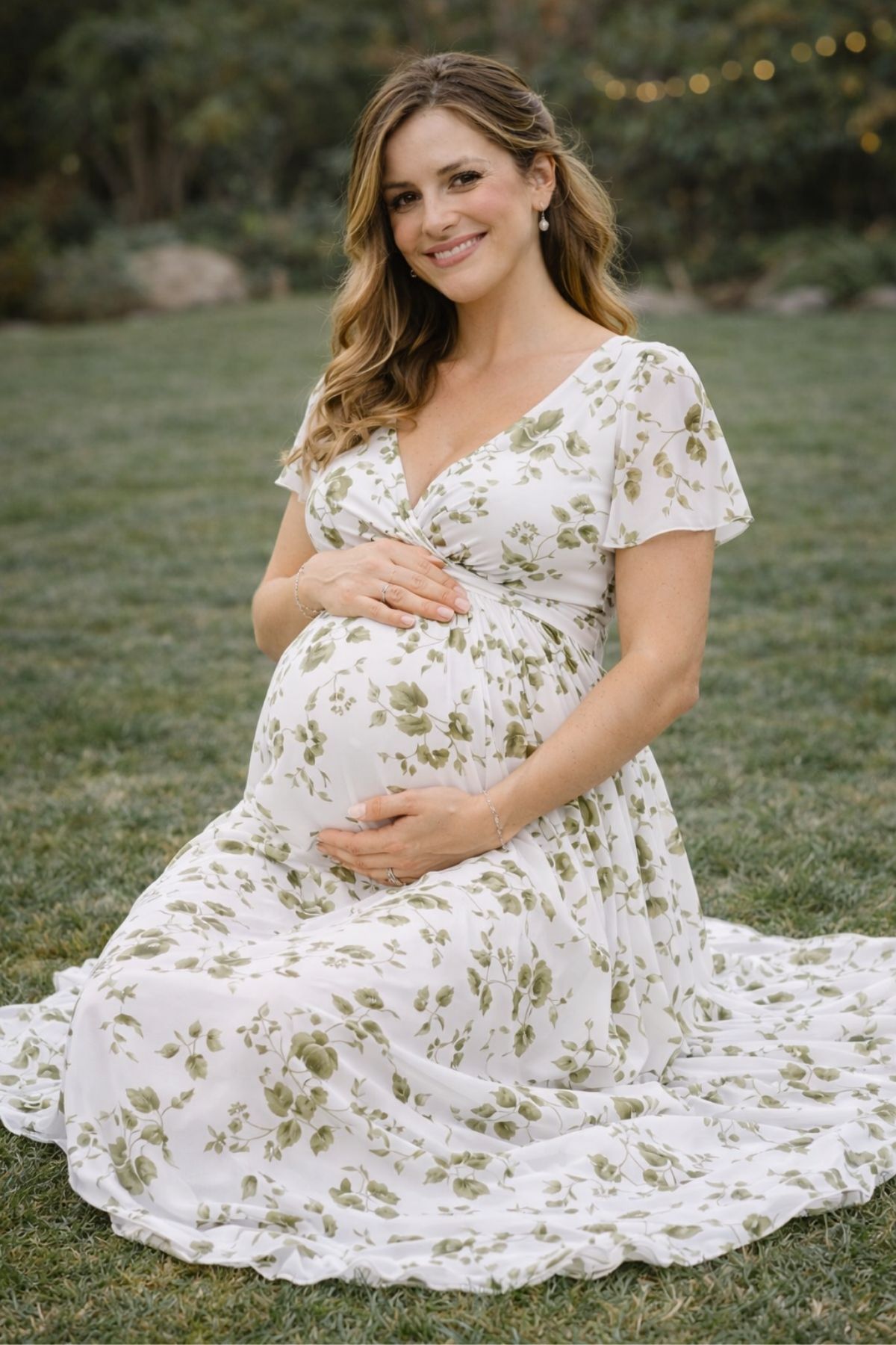 Pregnant bridesmaid in a soft green floral dress sitting on grass at an outdoor garden wedding.