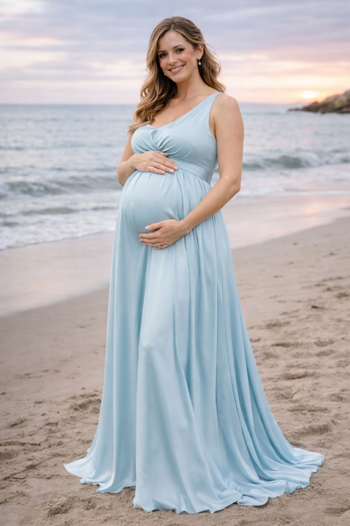Pregnant bridesmaid in a light blue one-shoulder dress standing by the beach at sunset.
