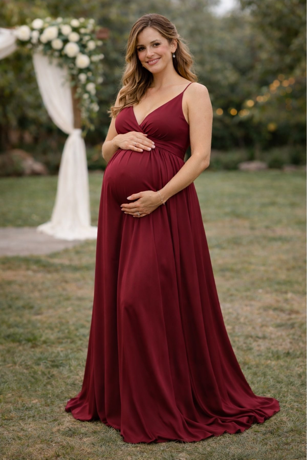 Pregnant bridesmaid wearing a deep burgundy flowing dress with thin straps at a garden wedding ceremony.
