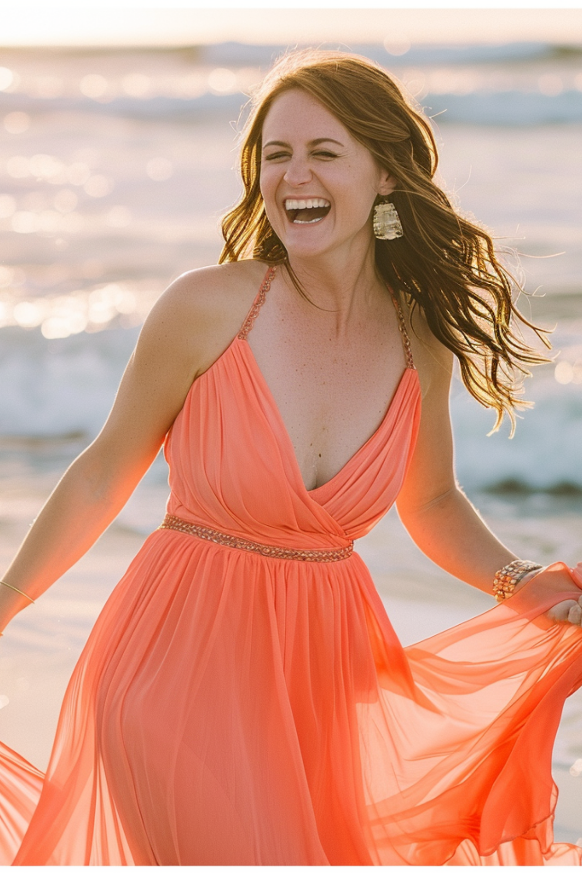 Woman wearing coral chiffon beach wedding guest dress laughing by the ocean during summer seaside wedding.