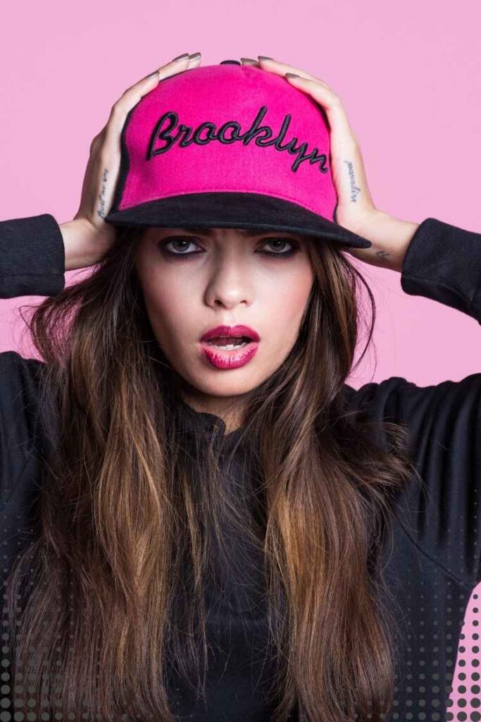 Close-up portrait of a confident woman wearing a pink snapback cap, black sweatshirt, and bold makeup. Modern Rebel Woman style combining streetwear fashion, individuality, confidence, and creative self-expression.