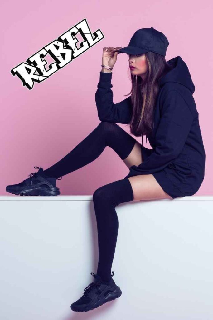 Young woman sitting against a pink background wearing an oversized black hoodie, black baseball cap, knee-high socks, and black sneakers, showcasing a bold urban streetwear style with a rebellious attitude.