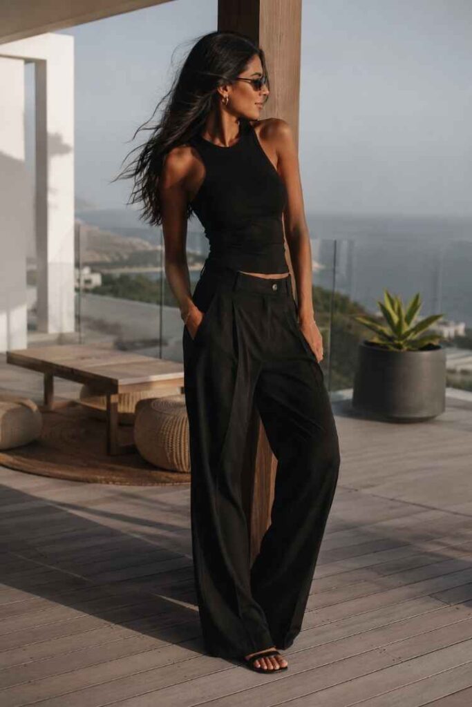 Elegant woman wearing a black sleeveless top and flowing black wide-leg trousers on a luxury terrace, creating a sophisticated minimalist summer look.